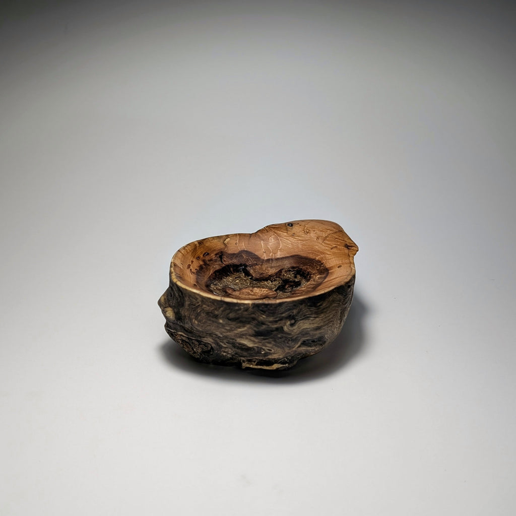 A small rustic wooden bowl with a rough, bark-like exterior and a smooth, hollowed interior, shown on a neutral gray gradient background.